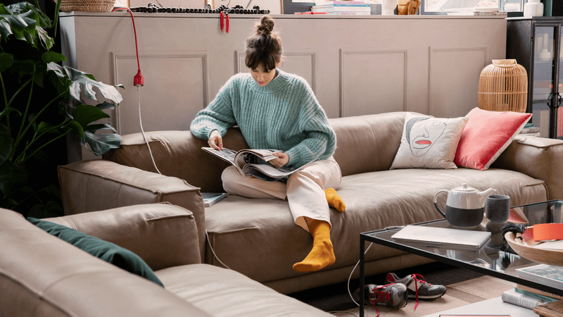 Eine Frau mit türkisfarbenem Pullover sitzt auf einem beigen Ledersofa in einem Wohnzimmer und liest eine Zeitschrift. Um sie herum sind bunte Kissen, eine Teekanne auf dem Tisch und Pflanzen im Hintergrund.