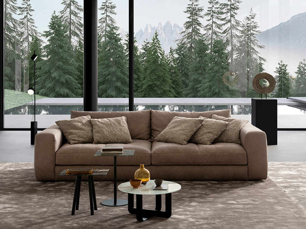Ein modernes Wohnzimmer mit einem braunen Ledersofa und mehreren Kissen, einem runden Couchtisch und einem Beistelltisch, vor einer großen Fensterfront mit Blick auf einen Nadelwald und Berge.