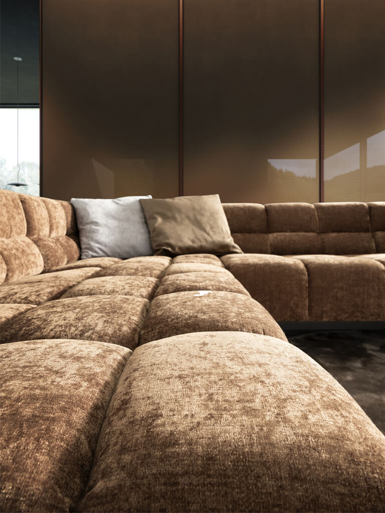 Eine Nahaufnahme des braunen, texturierten Stoffs eines modularen Sofas mit gestepptem Muster. Zwei Zierkissen, eines hellgrau und eines beige, liegen auf der Rückenlehne. Der Fokus liegt auf der Stoffstruktur und der Verarbeitung.