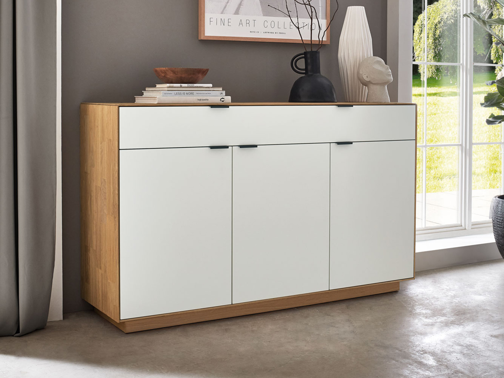 Wöstmann WM 2410 - Sideboard | Type 2550 | Lack und Wildeiche massiv Ein moderner Holzschrank mit weißer Front steht in einem stilvoll eingerichteten Wohnzimmer, dekoriert mit Büchern und Vasen.