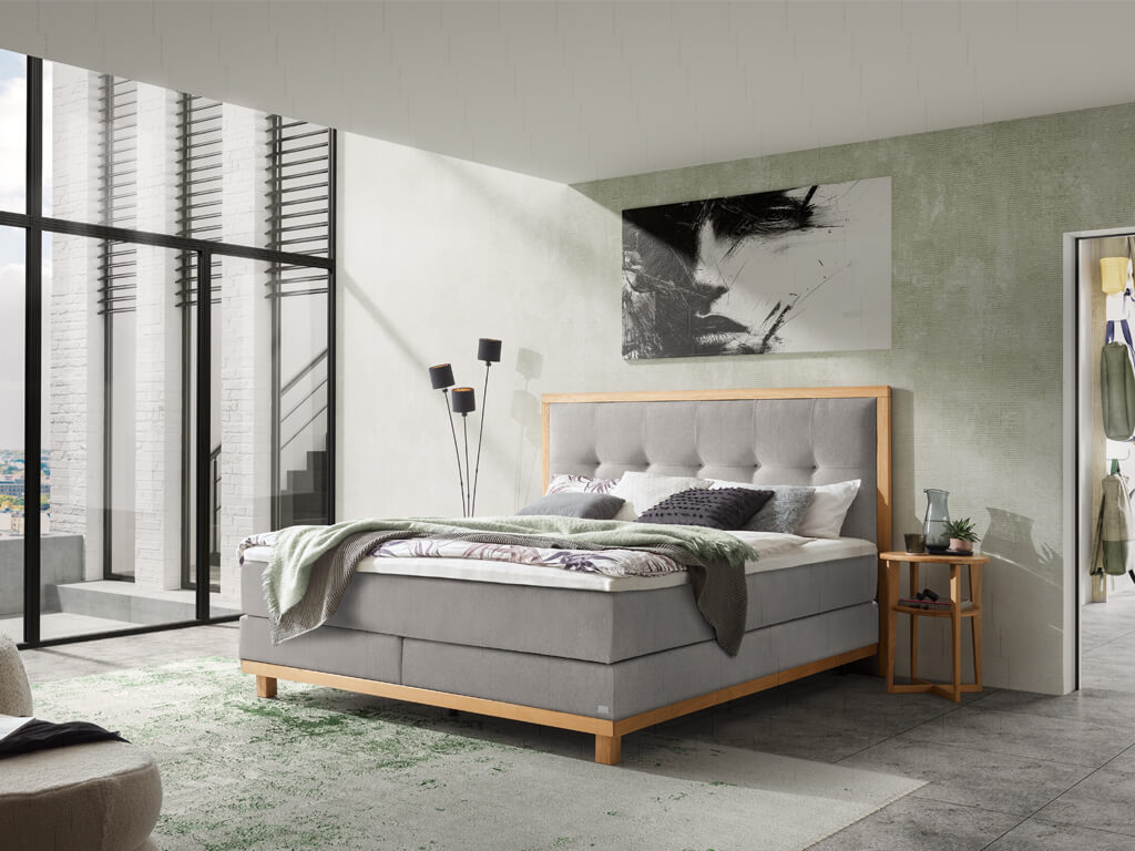 Ein modernes Schlafzimmer mit dem Hasena Boxspringbett "Pronto Cecilia", das einen hellgrauen Bezug und einen Holzrahmen hat, aufgenommen aus einem anderen Blickwinkel.