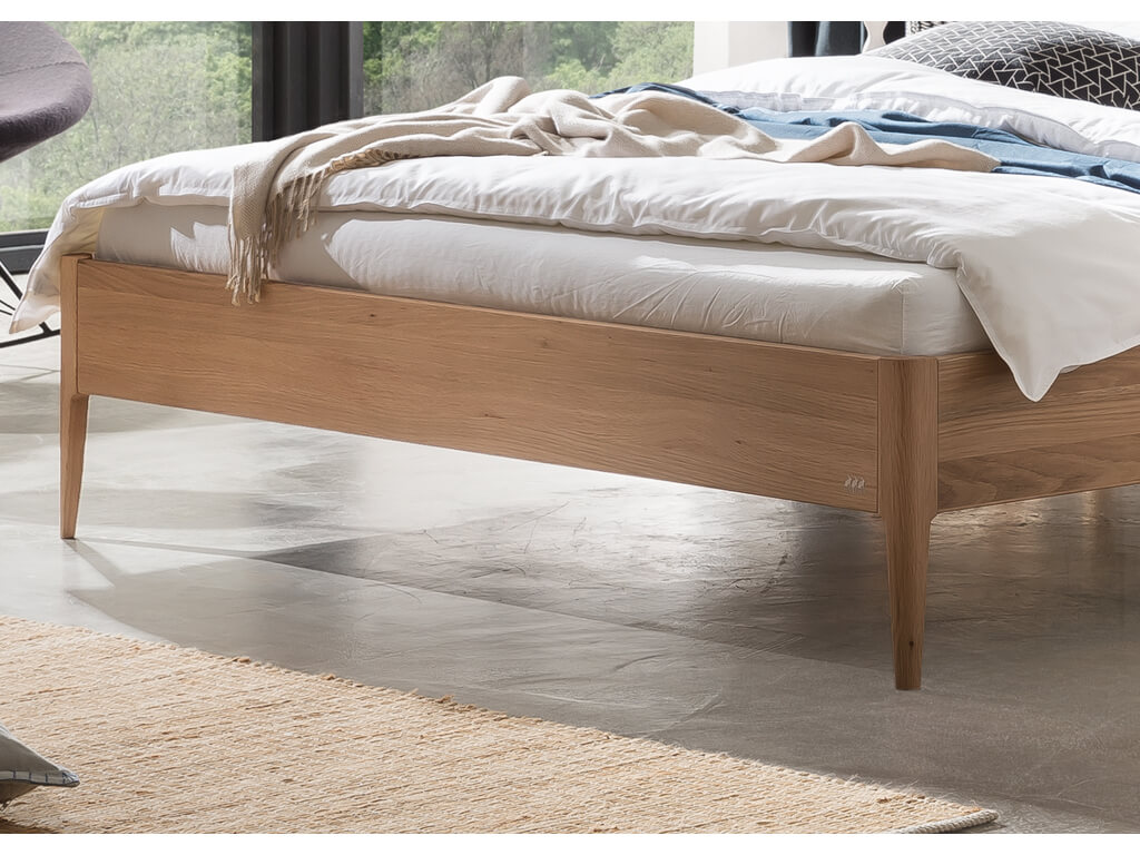 Nahaufnahme der Seite und der Füße eines Hasena Fanano Bettes. Der Bettrahmen ist aus hellem Eichenholz. Das Bett ist mit Bettwäsche gemacht und steht auf einem grauen Betonboden.