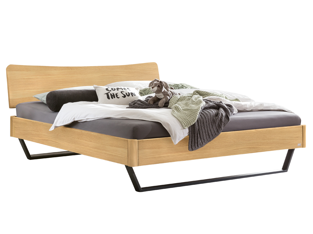 Ein Hasena Bettmodell Tresali aus hellem Eichenholz mit einem geschwungenen Kopfteil und schwarzen Metallfüßen, das mit Kissen und einer Decke dekoriert ist.