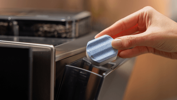 Eine Hand hält eine blau-weiße Siemens Entkalkungstablette über das geöffnete Pulverfach bzw. den Schacht eines Siemens EQ Kaffeevollautomaten, um den Entkalkungsvorgang zu starten.