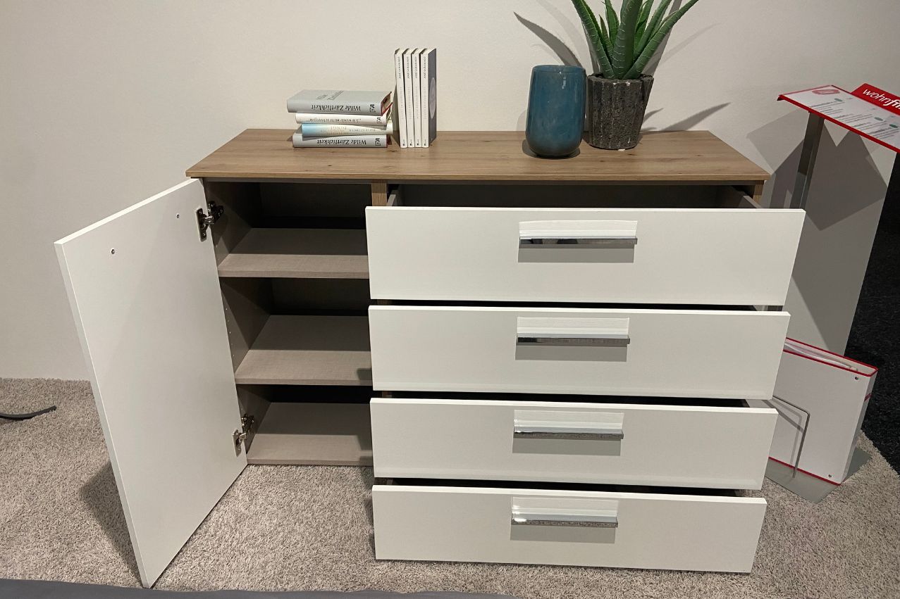 Moderner weißer Schrank mit vier geöffneten Schubladen, einer Tür, Büchern, einer Vase und einer Pflanze auf der Oberseite, auf Teppichboden stehend.