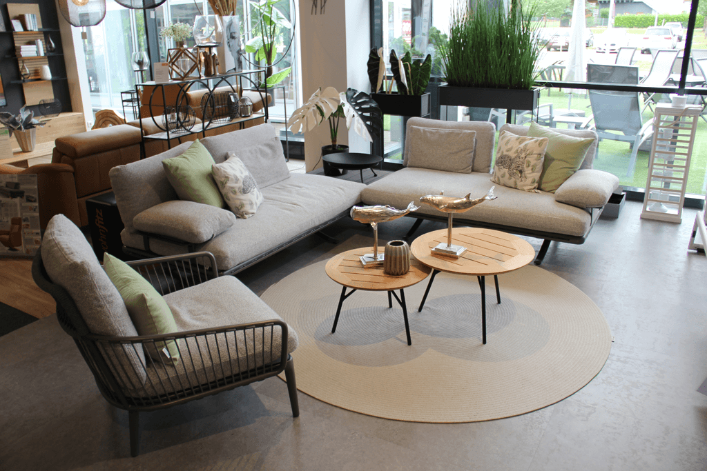 Rolf Benz  YOKO || Loungegruppe + Einzelsessel Eleganter Wohnbereich mit grauen Sofas, Kissen und dekorativen Tischen auf einem runden Teppich, umgeben von Pflanzen.