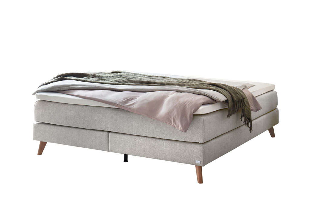 graues Boxspringbett mit braunen Holzfüße, stilvoll dekoriert mit Decken und Kissen vor weißem Hintergrund