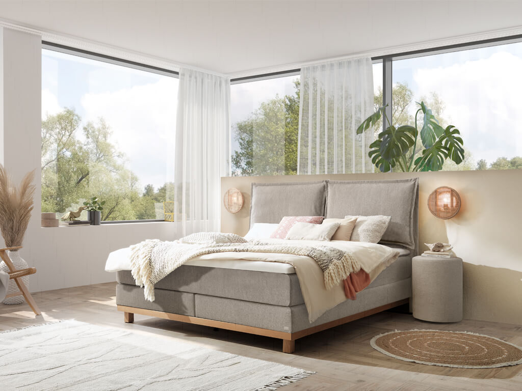 Ein Hasena Boxspringbett Pronto Zamora Dean mit hellgrauem Stoffbezug, das sich harmonisch in ein stilvolles, lichtdurchflutetes Schlafzimmer einfügt.