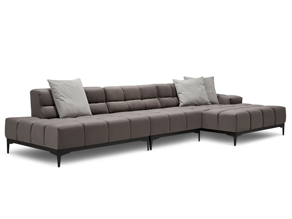 Ein langes L-förmiges Sofa in einem dunklen Taupe-Ton mit gesteppter Sitzfläche und Rückenlehne. Es steht auf schlanken schwarzen Metallfüßen. Zwei helle, graue Zierkissen liegen auf der Rückenlehne. Der Hintergrund ist weiß.