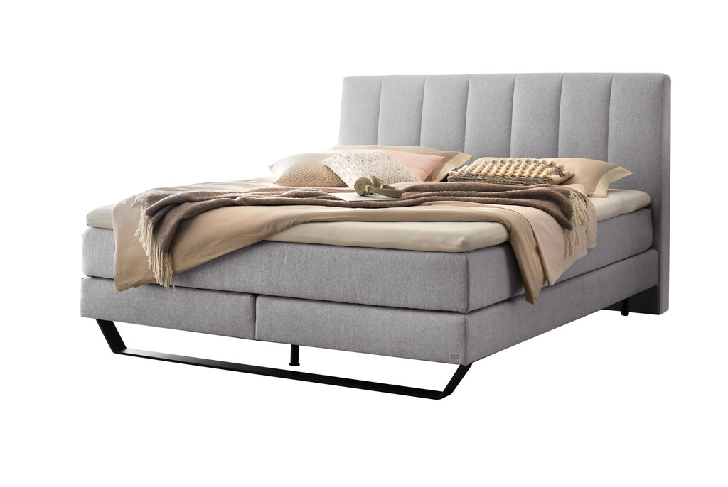 graues Boxspringbett mit Stahlkufe in schwarz, stilvoll dekoriert mit Decken und Kissen vor weißem Hintergrund