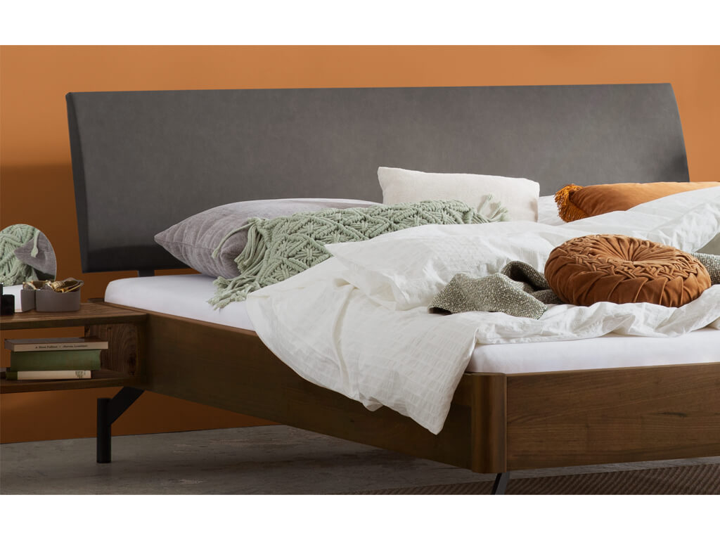 Ein Detailbild des Bettes "Broni" mit einem Rahmen aus Nussbaumholz. Das Bett verfügt über ein gepolstertes anthrazitfarbenes Kopfteil und dunkle Metallbeine. Daneben befindet sich ein Nachttisch aus Nussbaumholz, der am Bett befestigt zu sein scheint.