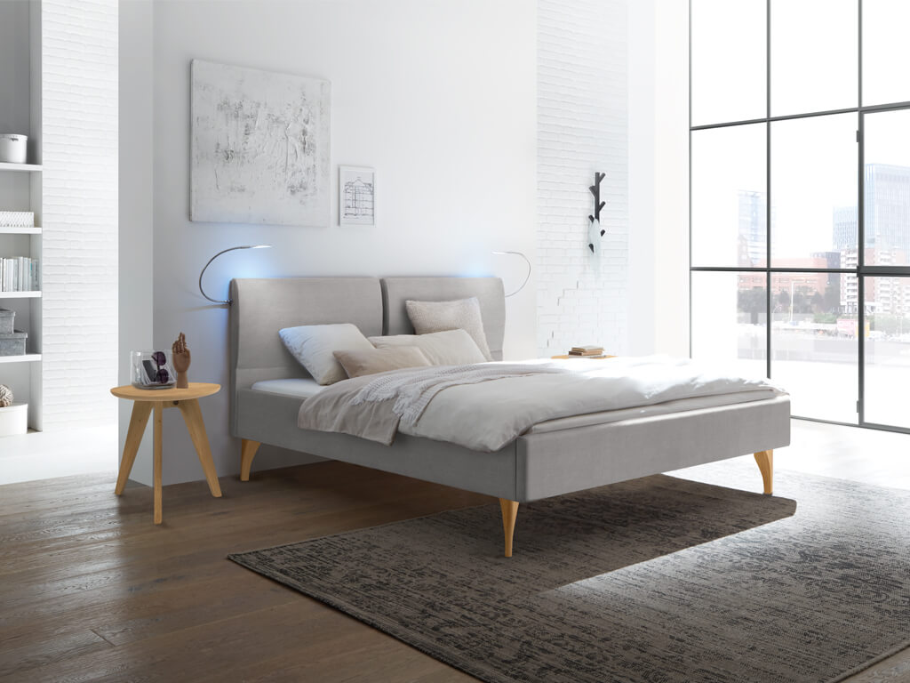 Ein graues Polsterbett mit geteiltem Kopfteil und hellen Holzfüßen in einem modernen Schlafzimmer mit großen Fenstern.