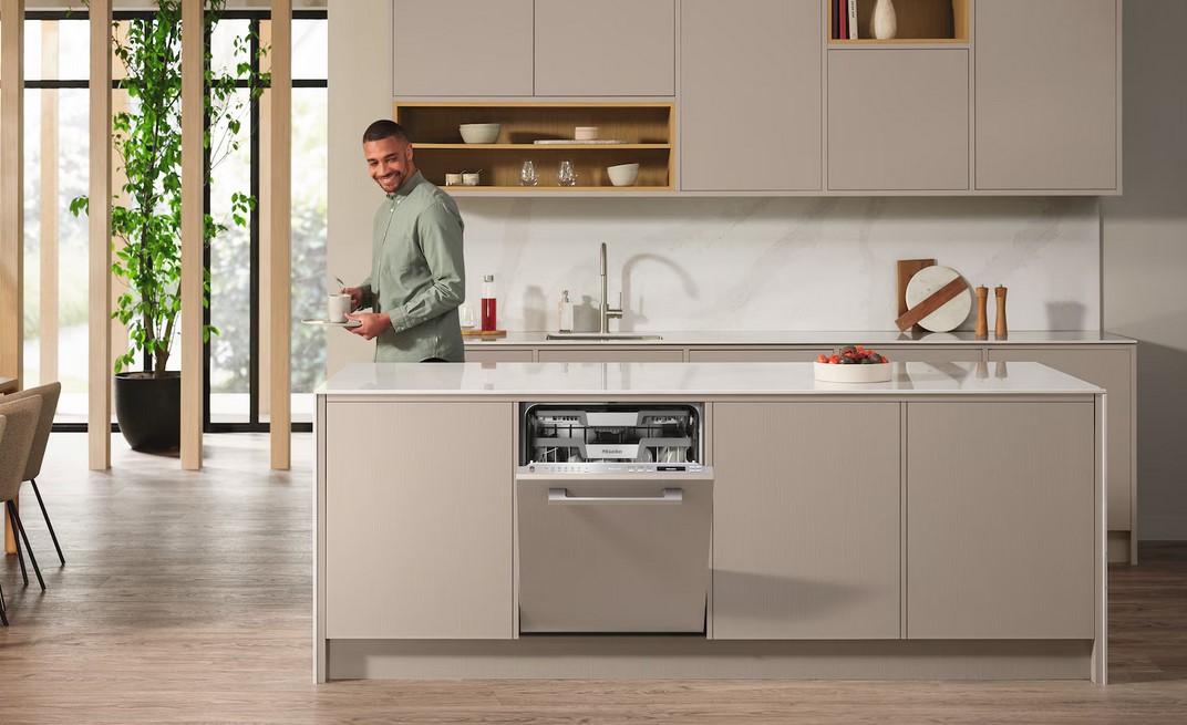 Ein lächelnder Mann steht in einer modernen Küche mit einem Kaffeebecher in der Hand, während im Vordergrund der Miele G 7191 SCVi AD Geschirrspüler in die Kücheninsel integriert ist und leicht geöffnet ist.