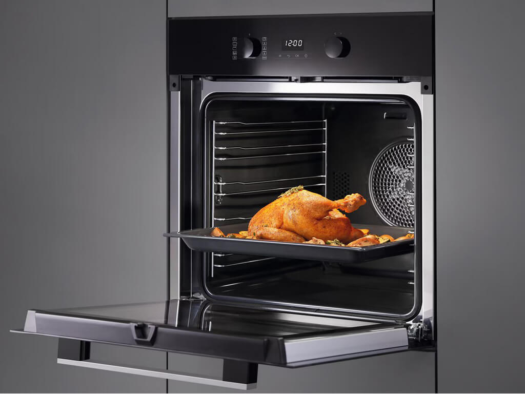 Eine Nahaufnahme des geöffneten Innenraums eines Miele-Backofens. Ein gebratenes, ganzes Huhn liegt zusammen mit Gemüse auf einem schwarzen Backblech, das auf einem ausziehbaren Rost in der Mitte des Ofens steht.