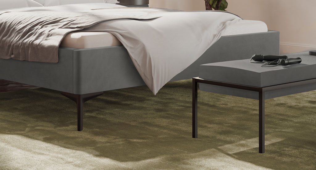 Nahaufnahme der schwarzen Bettfüße am grauen Polsterbett auf einem grünen Teppich im Schlafzimmer (KI-generiert)