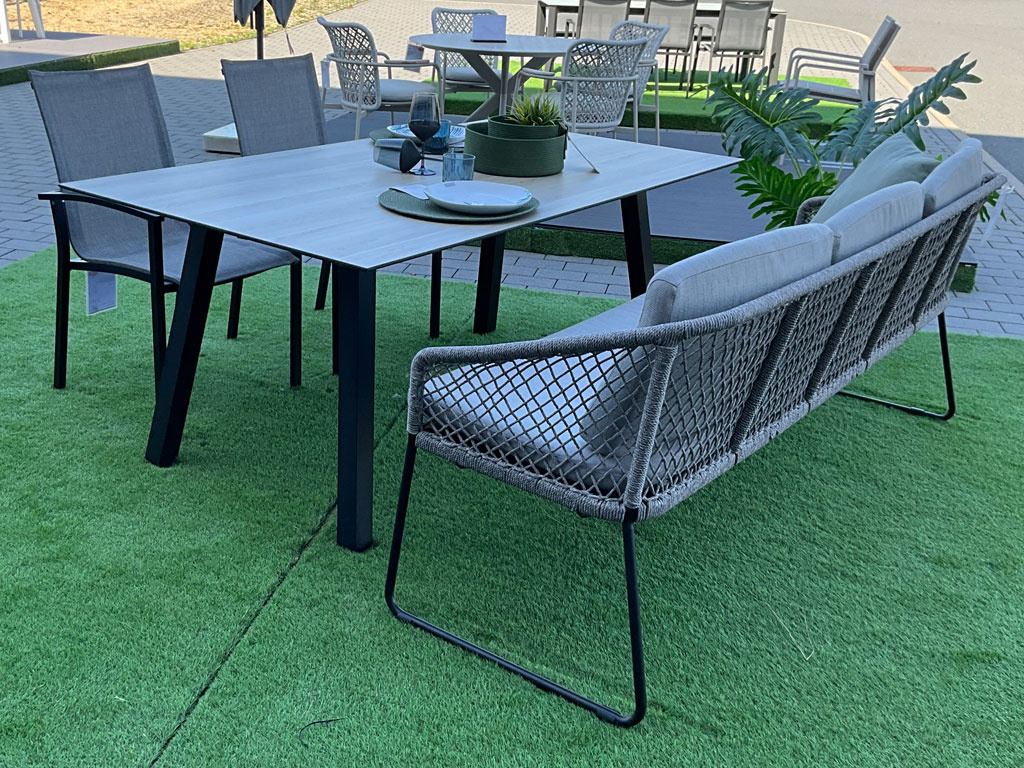 Moderne Garten-Sitzgruppe mit großem Esstisch, grauer Tischplatte, schwarzen Metallbeinen und mehreren grauen Gartenstühlen mit geflochtener Rückenlehne.