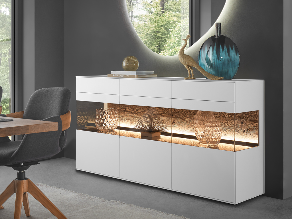 hartmann Torido - Sideboard | Lack weiß / Kerneiche Umato | B: 172,8 cm Ein modernes, weißes Hartmann Torido Sideboard 4171 mit beleuchteten Holzfächern und Glastüren, das in einem eleganten Ess- oder Wohnbereich steht. Dekorationsartikel wie Vasen und Skulpturen sind in den Fächern platziert.
