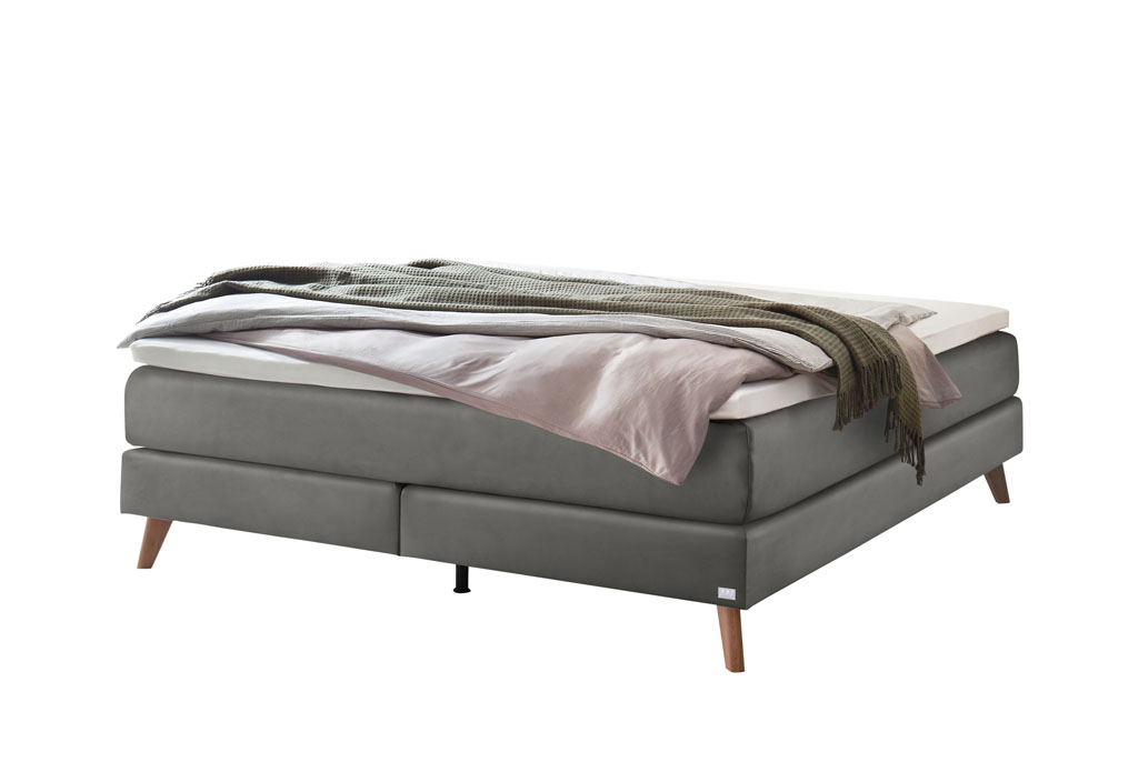 graues Boxspringbett mit braunen Holzfüße, stilvoll dekoriert mit Decken und Kissen vor weißem Hintergrund