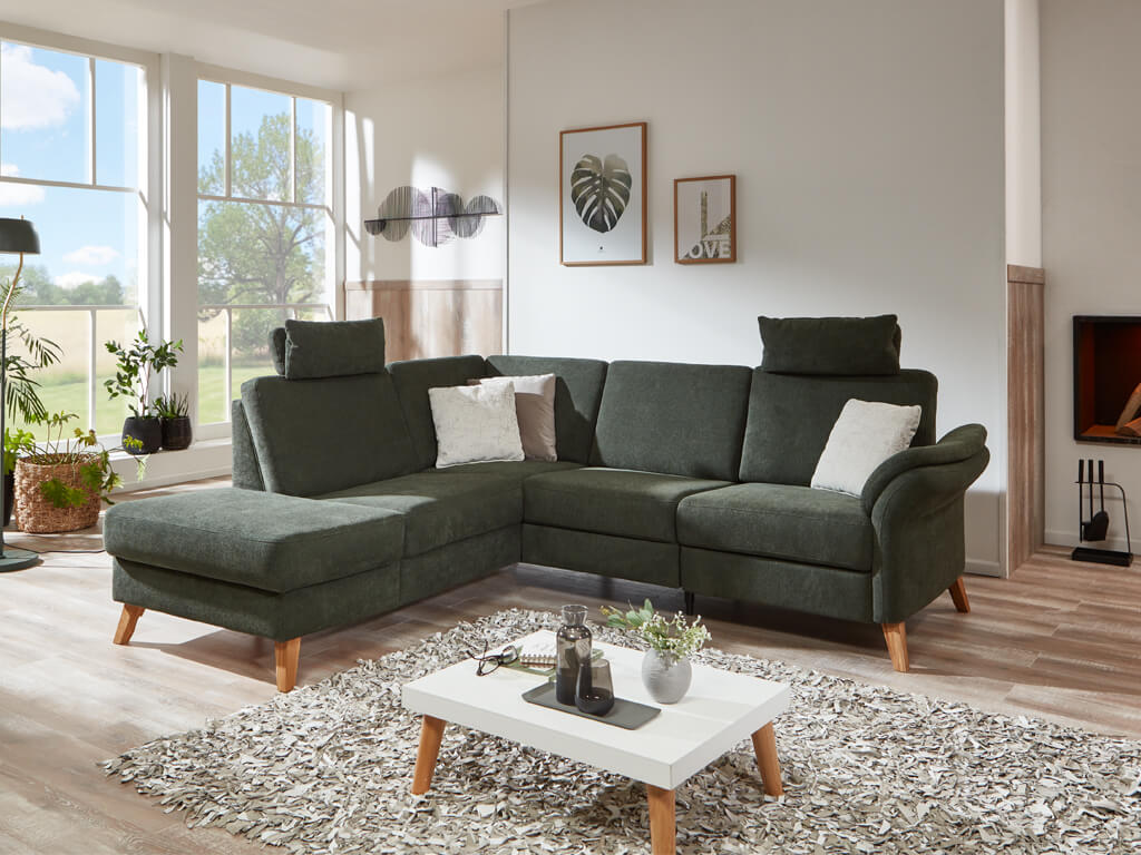 sit&more Freiburg | Polstergarnitur | 5729/5730 Dunkelgrünes Ecksofa mit weißen Kissen in einem hellen Wohnzimmer mit Holzboden und großen Fenstern.