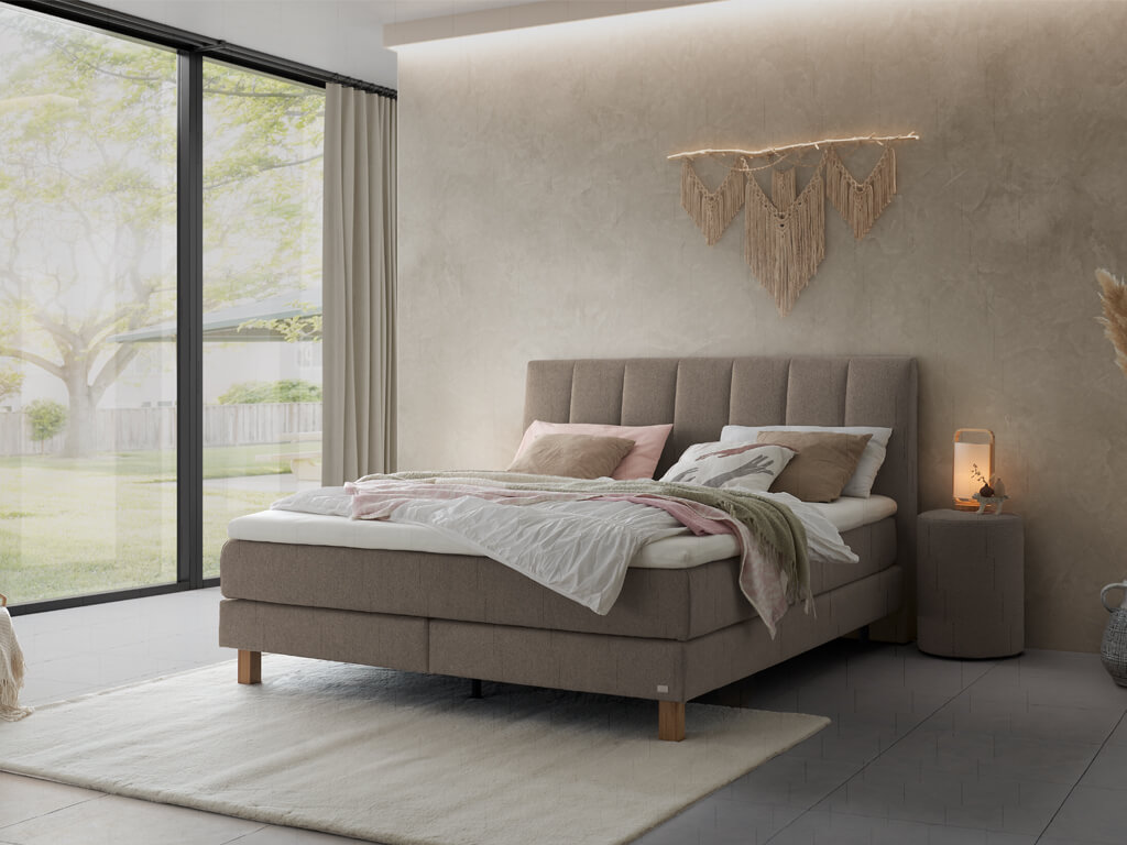 Ein Boxspringbett in dunklem Beige mit einem gesteppten Kopfteil und einem passenden Nachttisch in einem eleganten, minimalistischen Raum.