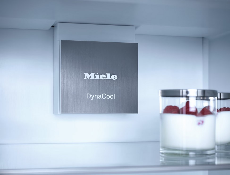 Nahaufnahme der Miele DynaCool-Lüftungseinheit aus gebürstetem Metall, die prominent im Inneren eines hell beleuchteten Miele K 7748 D Kühlschranks angebracht ist. Im Vordergrund sind zwei Gläser mit Joghurt und Himbeeren auf einem Regal zu sehen.