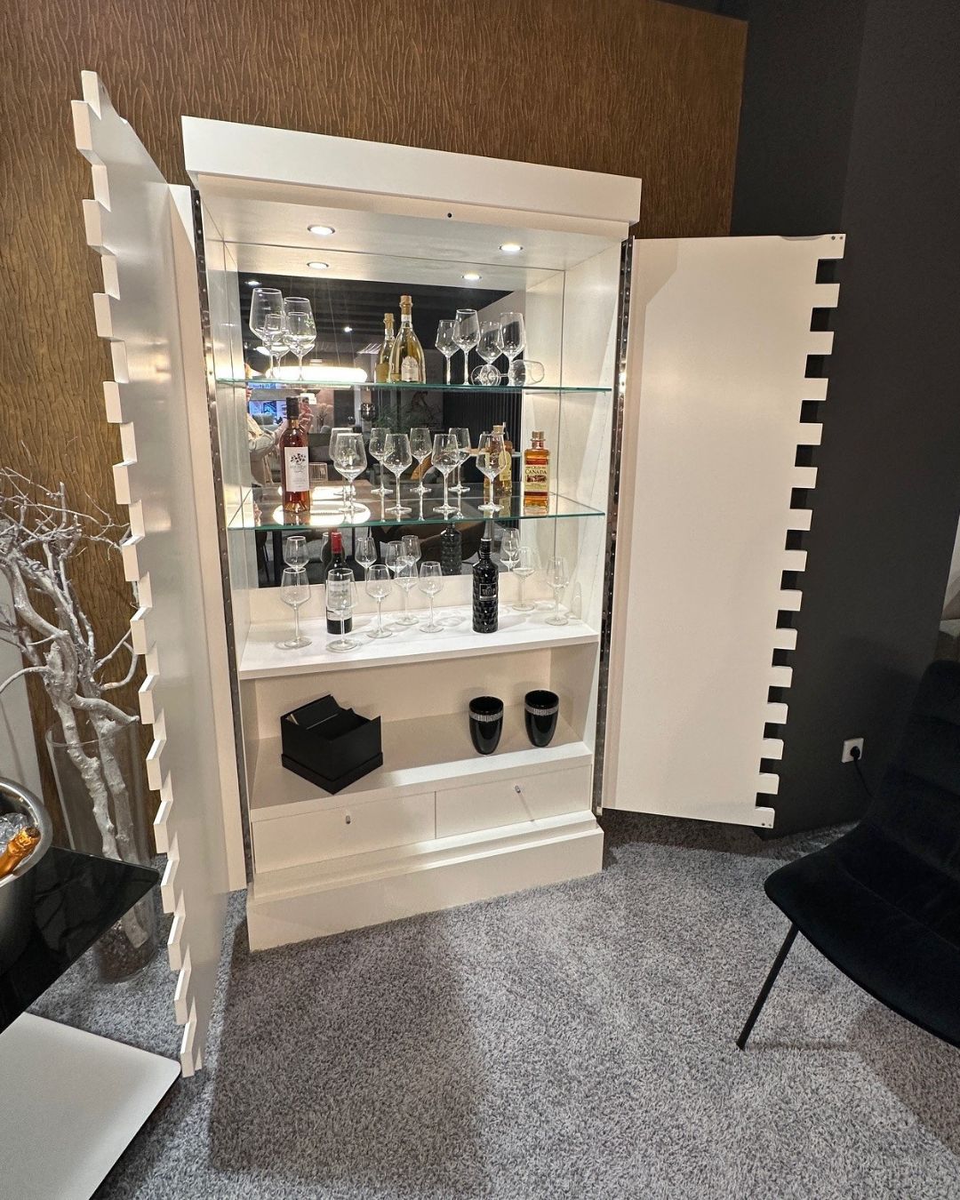 KMK MAXIM | Barschrank Ein eleganter weißer Vitrinenschrank, der eine Auswahl an Weingläsern und verschiedenen Getränkeflaschen präsentiert, in einem stilvoll eingerichteten Raum.