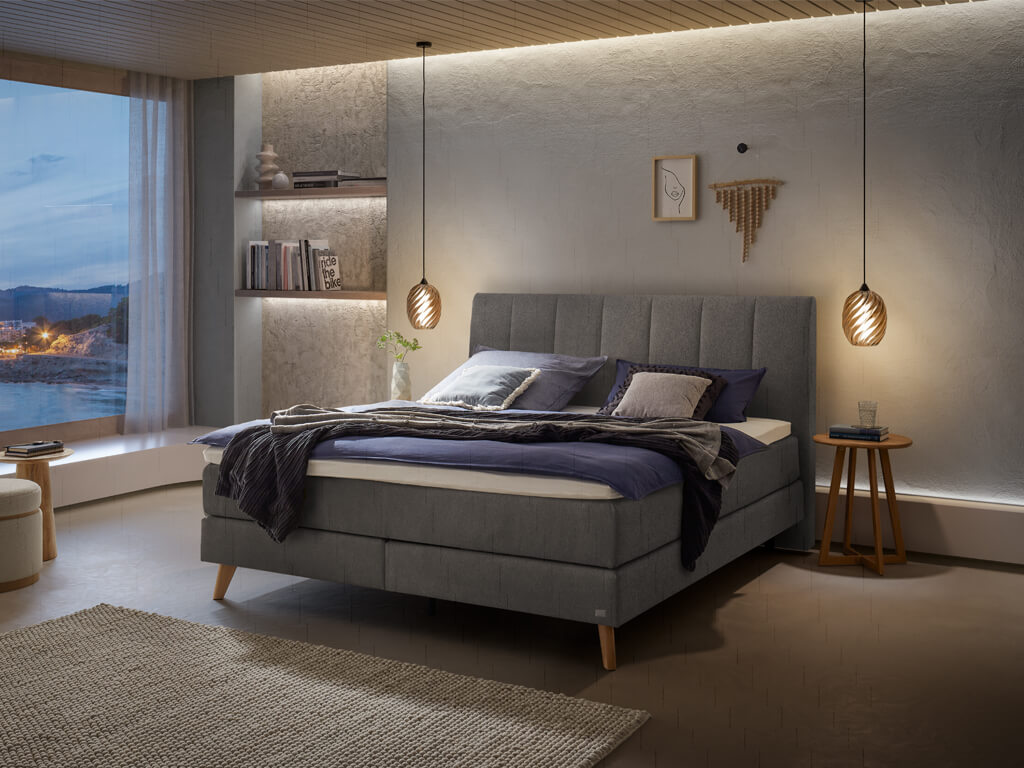 Ein Boxspringbett in grauer Stoffpolsterung und Holzfüßen in einem modern gestalteten Schlafzimmer bei Nacht.