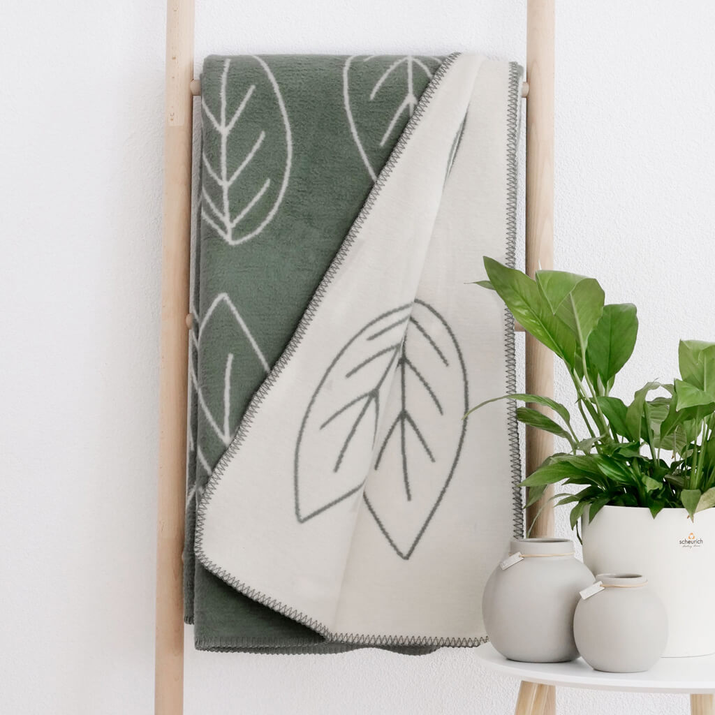 RIEMA. BENTE – Wohndecke, Bio-Baumwolle - Blätter | 150 x 200 cm | grün-natur Grüne und weiße Kuscheldecke mit Blätterdesign auf Holzleiter neben grüner Pflanze