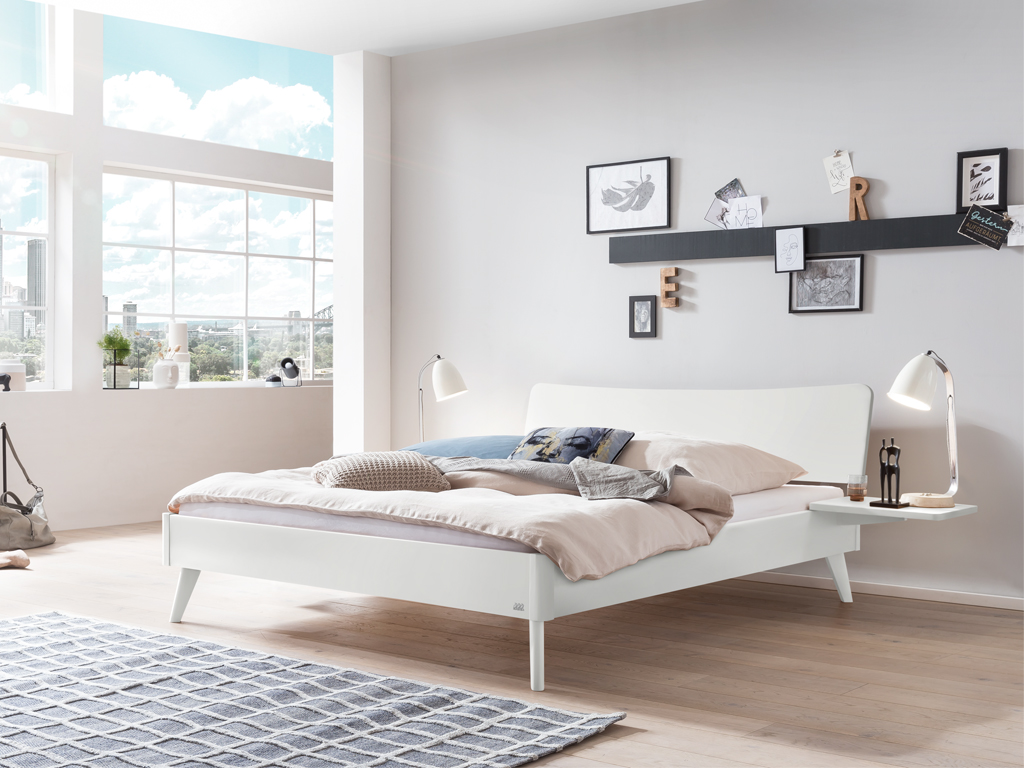 Ein weiß lackiertes Hasena Bett Caluso in einem hellen, modern eingerichteten Schlafzimmer. Das Bett hat ein geschwungenes Kopfteil und schräg ausgestellte Beine.