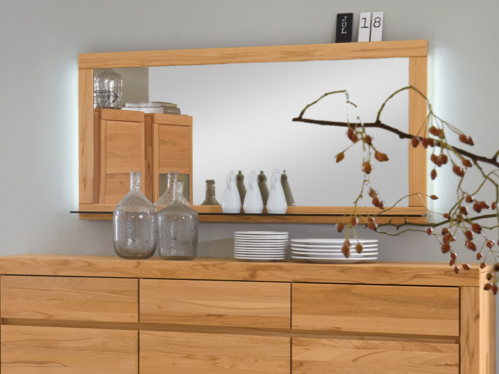 Ein großer, rechteckiger Spiegel in einem Holzrahmen hängt über einer Holzkommode. Auf der Kommode stehen dekorative Glasvasen und gestapelte Teller.