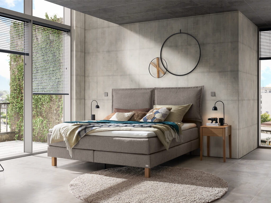 Ein modernes Schlafzimmer mit einem grauen Hasena Boxspringbett "Pronto Bonita" und einem Beton-Look an den Wänden.