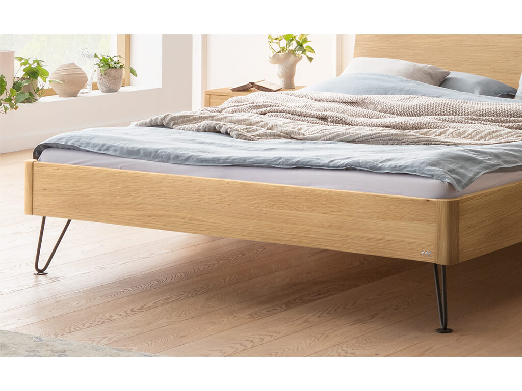 Nahaufnahme des unteren Teils eines Hasena Bett Locarno mit einem Rahmen in heller Eiche (Eiche bright) und schwarzen, anthrazitfarbenen Haarnadel-Beinen. Man sieht den Bettrahmen, die Beine und den Holzboden.