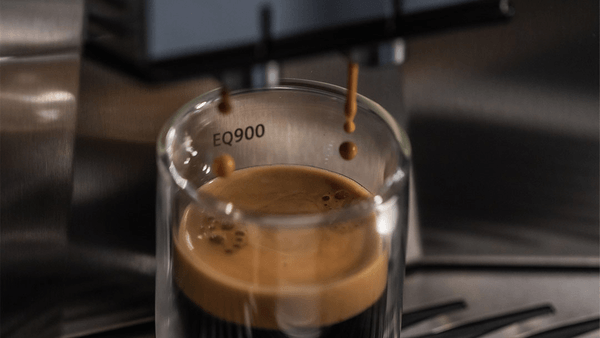 Frisch gebrühter Espresso mit einer feinen, goldbraunen Crema fließt aus dem Auslauf eines Siemens EQ900 Kaffeevollautomaten in ein transparentes, doppelwandiges Glas.