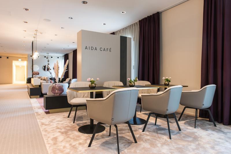 Elegantes Interieur eines Cafés mit hellen Sesseln, zeitgemäßer Dekoration und lila Vorhängen, beschriftet mit 'Aida Café'.