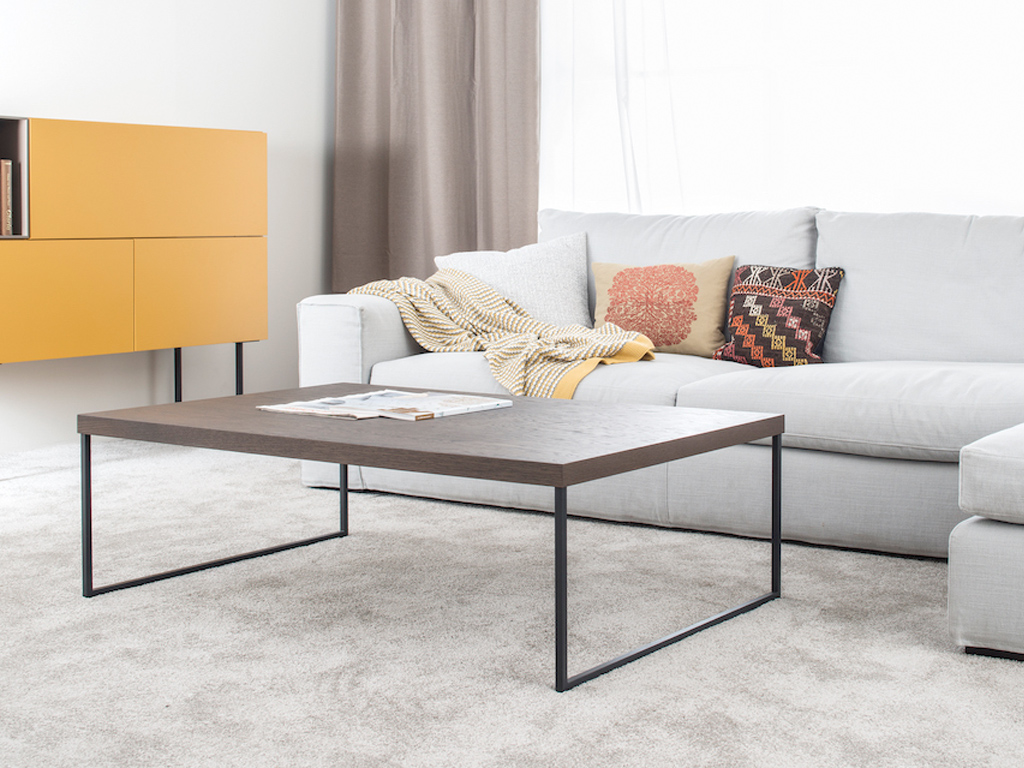 Sudbrock Cubo - Couchtisch | B: 120 cm | Wildeiche coffee furniert Geräumiges Wohnzimmer mit grauem Sofa, dekorativen Kissen, einem schlichten Couchtisch aus Holz und Metall sowie einer gelben Kommode.