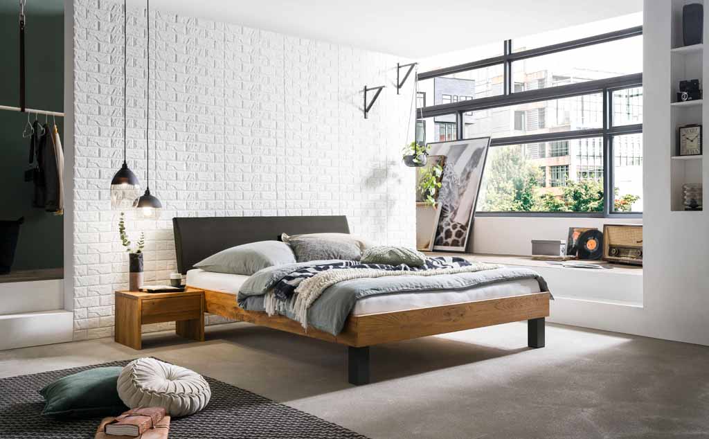 Helles und modernes Schlafzimmer mit einem Holzrahmenbett, weißen Ziegelwänden, großen Fenstern und stilvollen Dekorationen. Neben dem Bett steht ein kleiner Holztisch mit einer Lampe.