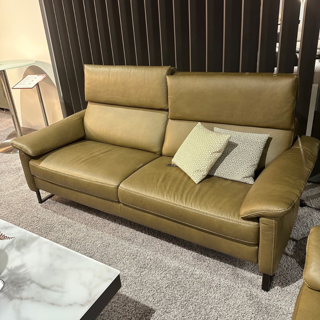 Eine moderne olivgrüne Ledercouch mit zwei gemusterten Kissen in einem stilvollen Wohnzimmer auf grauem Teppichboden neben einem Marmortisch.