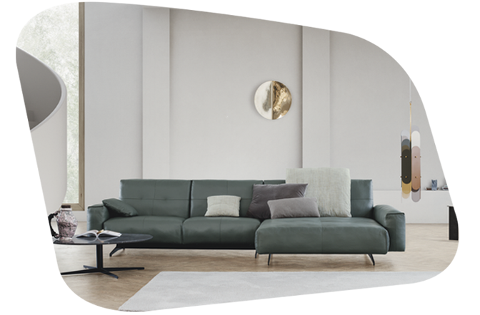 Im Zentrum steht ein großes, L-förmiges dunkelgrünes Sofa mit Kissen, das auf einem weißen Teppich platziert ist. Vor dem Sofa befindet sich ein kleiner runder Couchtisch mit Büchern und Dekorationsgegenständen darauf.