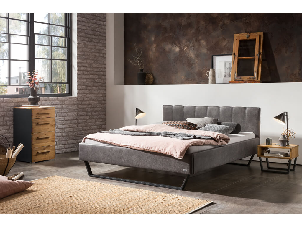 Ein modernes Schlafzimmer mit einem dunkelgrauen Polsterbett, einem Nachttisch und einer Kommode in einer industriell wirkenden Umgebung.