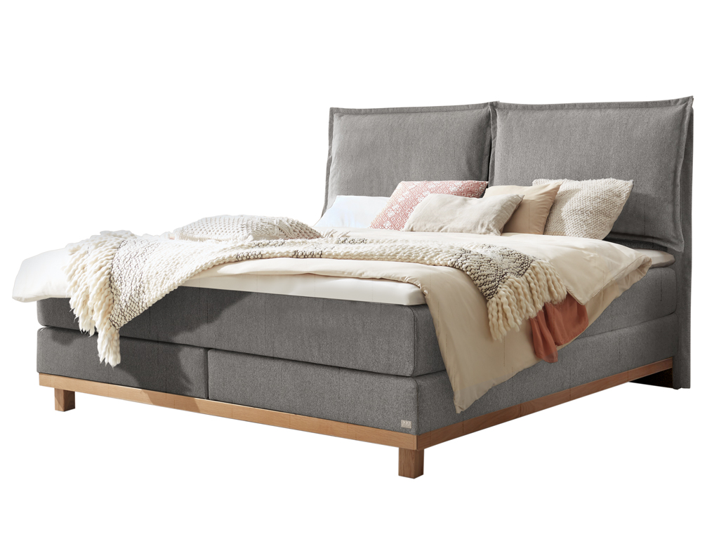 Freigestelltes, graues Boxspringbett mit hellem Holzbettgestell und hoher, zweiteiliger Lehne.