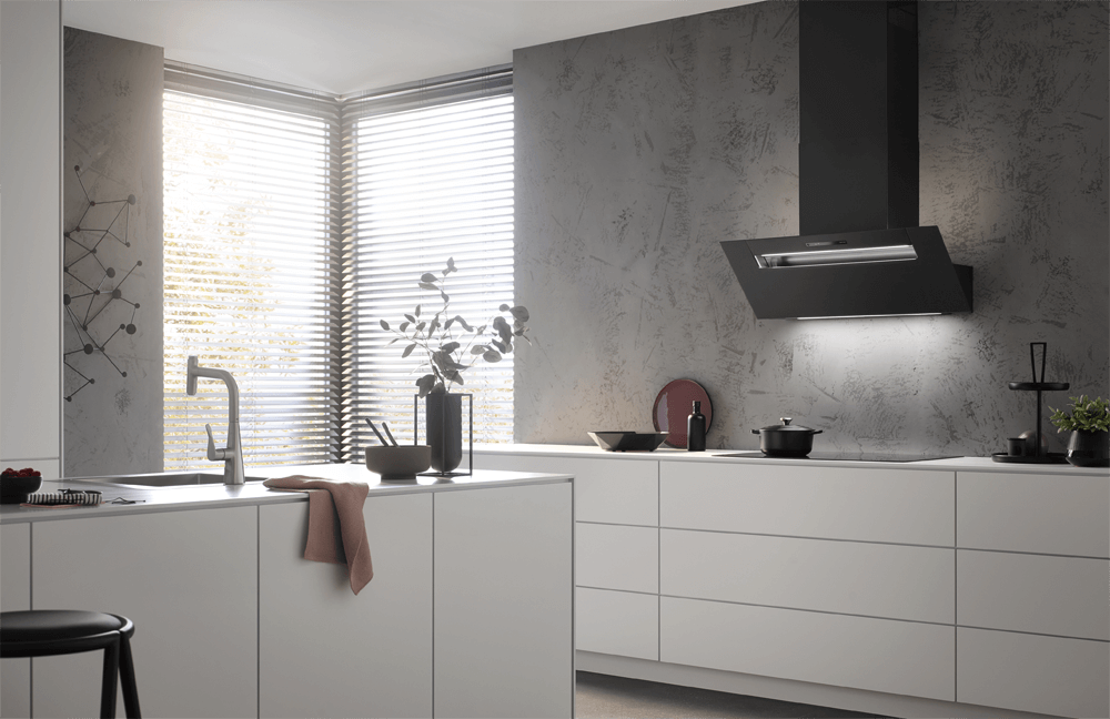 Eine stilvolle Küche hat eine mattschwarze schräge Dunstabzugshaube als Blickfang, die mit einer strukturierten grauen Wand kontrastiert. Die weißen Schränke und der helle Holzboden schaffen einen modernen und einladenden Raum mit natürlichem Licht.