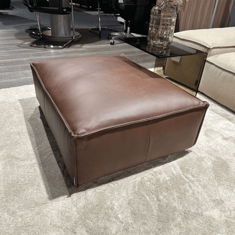 designwerk PUZZLE | Hocker Ein brauner Lederhocker auf einem beigen Teppich, ideal für moderne Wohnzimmer oder Büros.