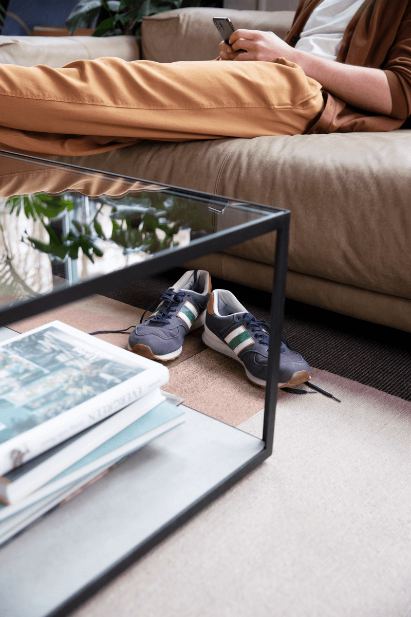Ein Paar blaue Sneakers mit weißen und grünen Streifen steht neben einer Couch. Eine Person liegt auf dem Sofa und benutzt ein Smartphone.
