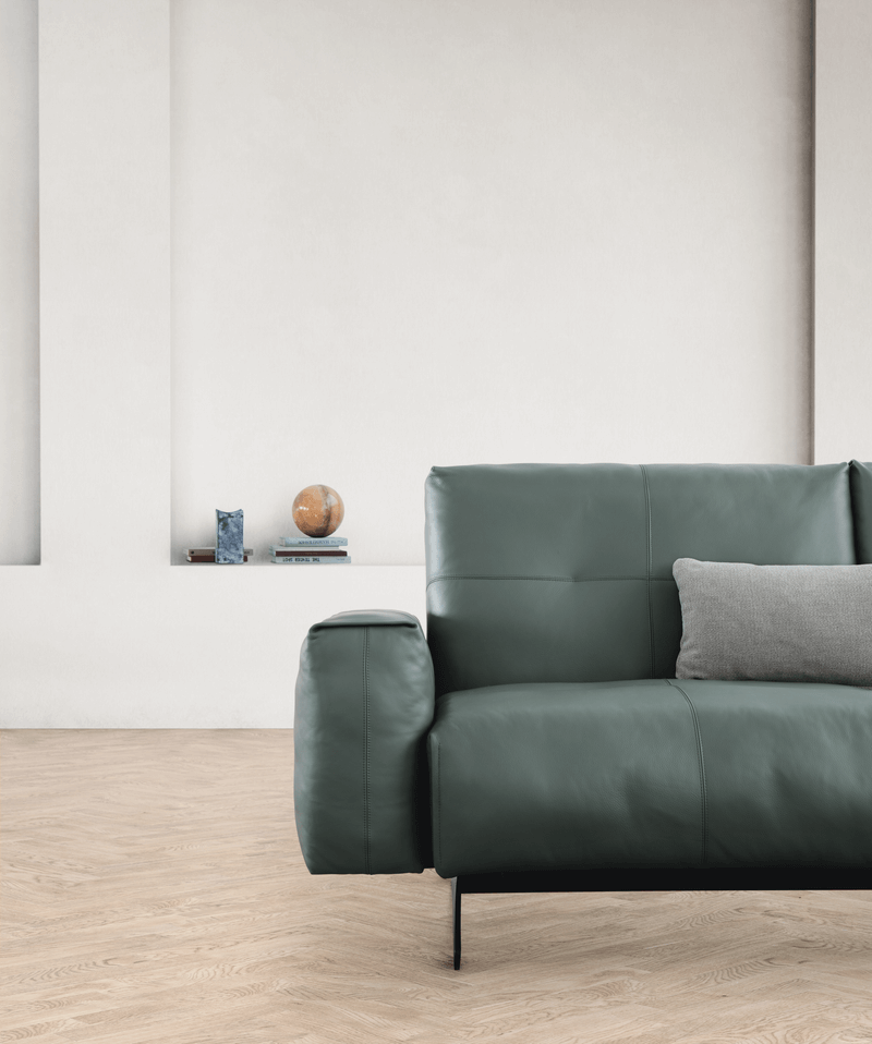 Das Bild zeigt ein minimalistisches Wohnzimmer mit einem grünen Ledersofa von Rolf Benz 50, zwei Kissen und einem kleinen Bücherstapel auf dem Boden vor einer weißen Wand.
