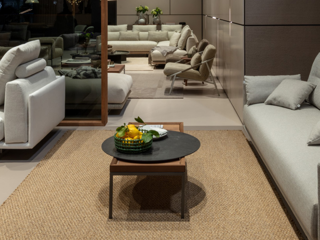 rechteckiger brauner Couchtisch mit schwarzem Gestell und einer runden aufliegenden Platte stilvolldekoriert in einem Ausstellungsraum mit vielen grauen Sofagarnituren und Sessel