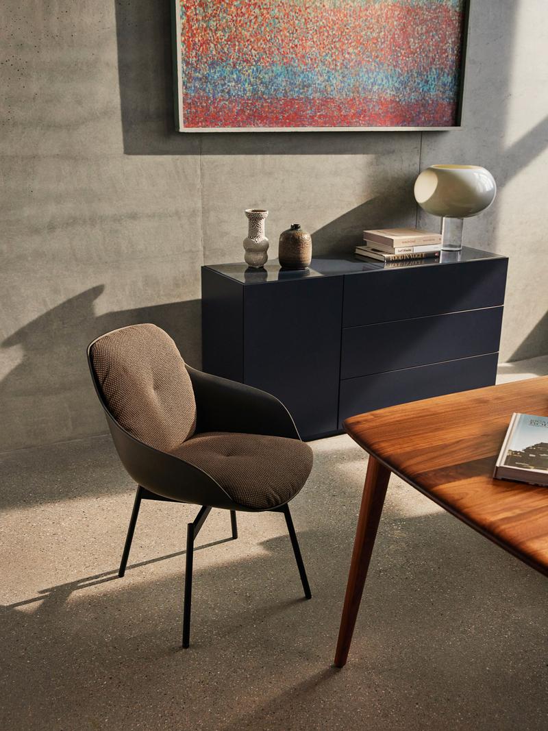 Ein eleganter Esszimmerstuhl neben einem Holztisch, im Hintergrund eine blaue Kommode mit Dekoration und ein abstraktes Kunstwerk an der Wand.