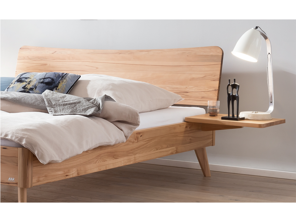 Ein hölzernes Hasena Bett Caluso aus Wildbuche. Das Bett hat ein geschwungenes Kopfteil und schräg ausgestellte Beine. An der Seite ist ein schwebender Nachttisch mit einer Lampe und einer kleinen Skulptur zu sehen.