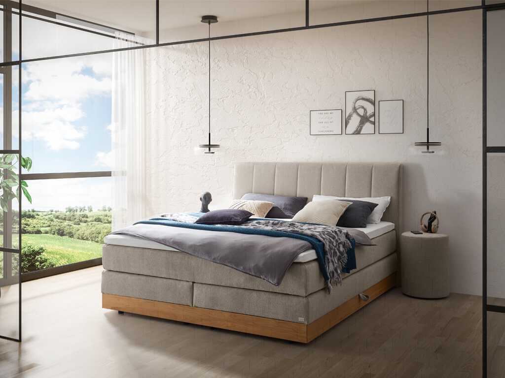 Ein Schlafzimmer mit einem hellgrauen Boxspringbett, das von hellen, strukturierten Wänden und großen Fenstern umgeben ist.