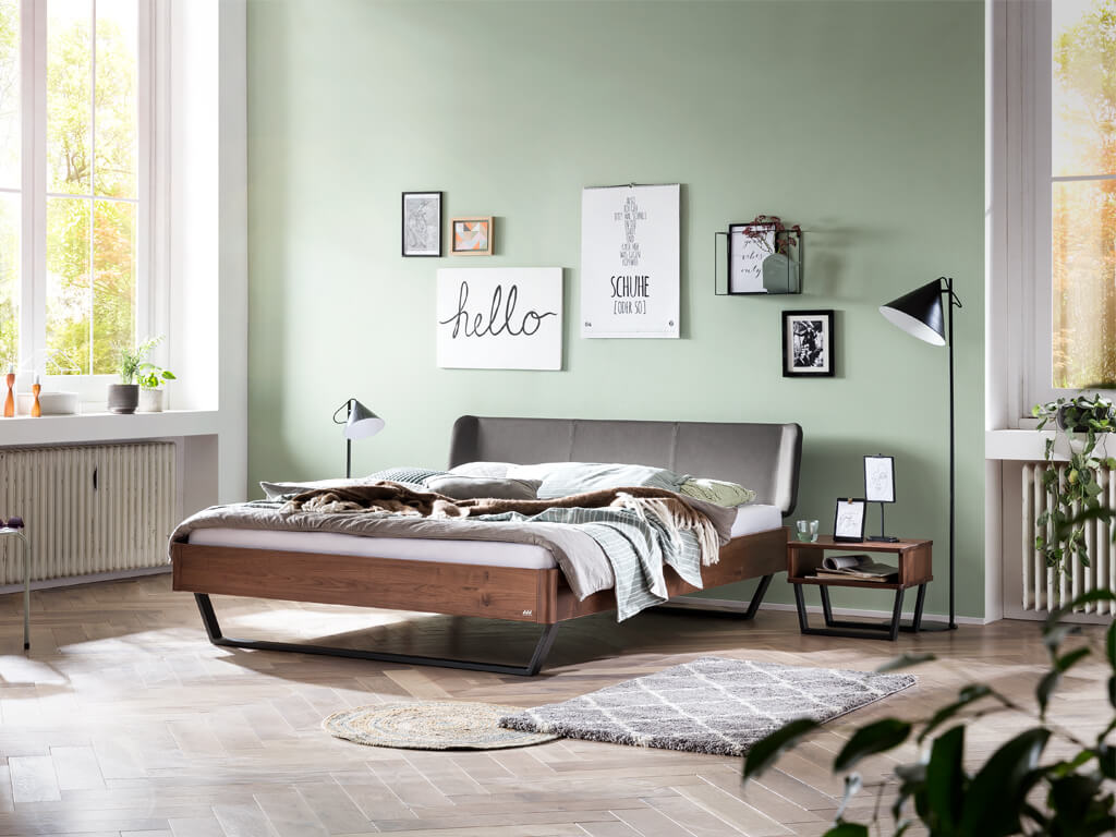 Hasena -  Lounge Bett Montieri | Polsterkopfteil Boga | Metallfuß Glyn | versch. Größen & Farben konfigurierbar Ein Schlafzimmer mit einem Holzbett aus dunkelbraunem Nussbaum. Das gepolsterte Kopfteil ist in Anthrazit gehalten. Das Zimmer hat mintgrüne Wände und ist mit Bildern, einer Stehlampe und einem passenden Nachttisch in Nussbaum dekoriert.