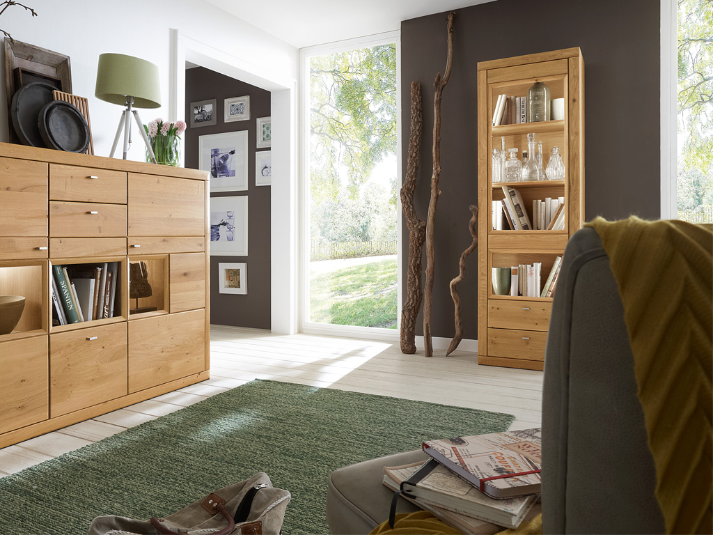 Ein helles Wohnzimmer mit modernen Holzmöbeln. Links steht ein Sideboard mit Ladeflächen und Regalen, dekoriert mit Lampen und Pflanzen. Rechts sieht man ein hohes Bücherregal vor einer dunkelgrauen Wand. Durch die große Fensterfront fällt viel Tageslicht